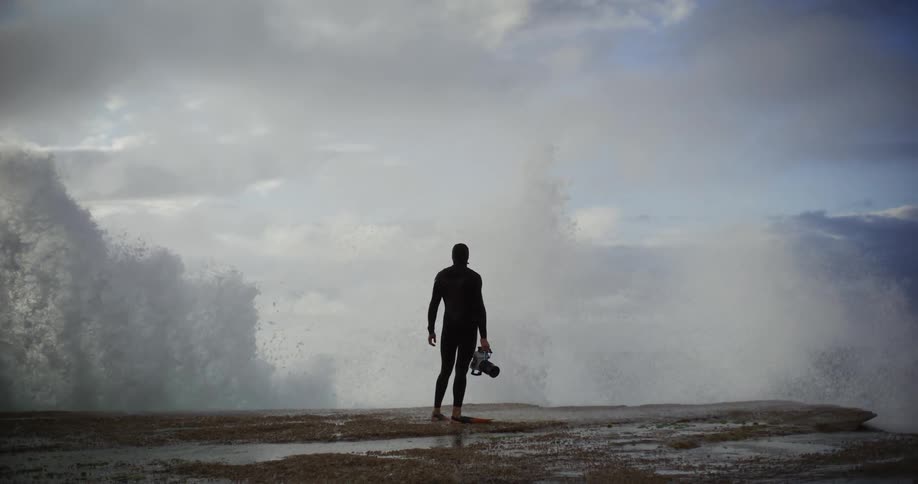 Man wearing scuba diving gear and holding a camera on his hand next to the shoreline with a wave breaking against the rocks outdoors during day time