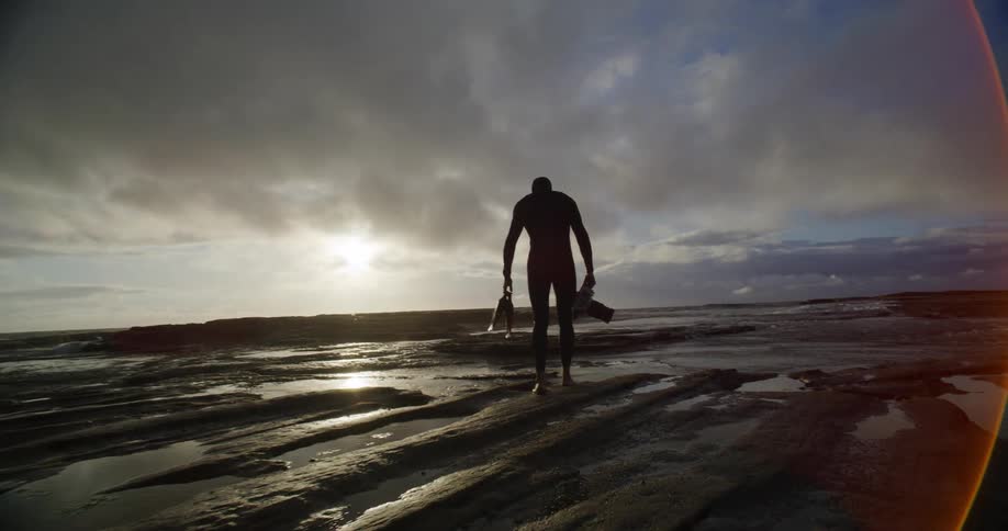 Man wearing scuba diving gear walking outdoors on a beach carrying a camera in his hand during dusk or dawn