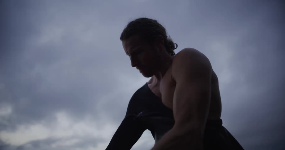 A young muscular man wears a black wetsuit on the coast under an overcast sky
