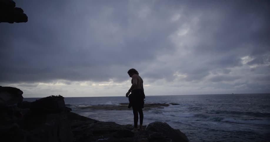 A man is standing on a rocky beach as he takes his shirt off