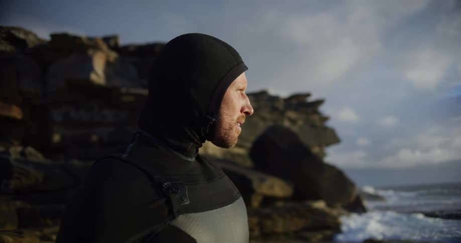 Bearded man wearing scuba diving clothes standing on a beach sighing and smiling outdoors during day time