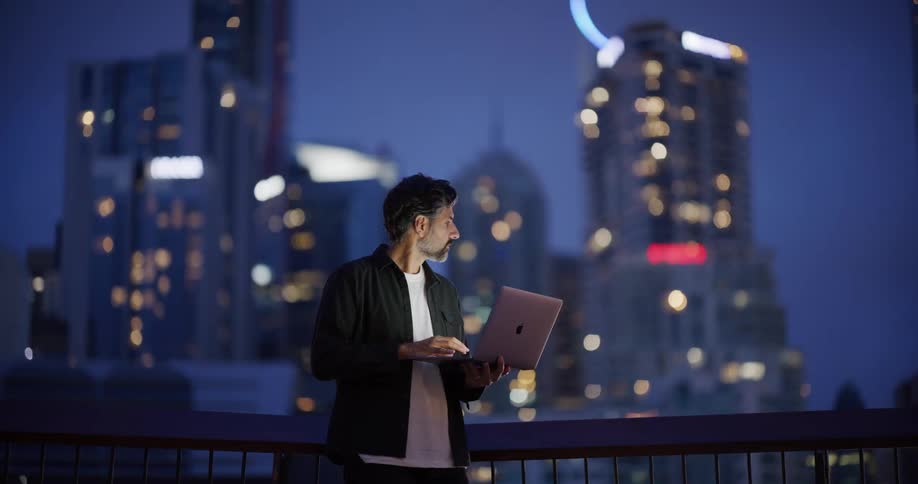 Man standing in an outdoor area at night typing on a laptop