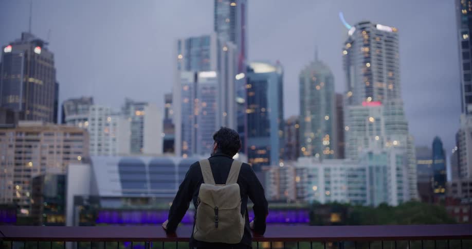 A person wearing a backpack looks out at a city skyline at dusk