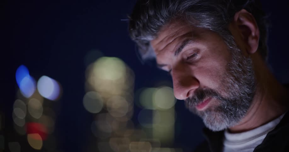 A man with gray hair and a beard looking down with blurred city lights in the background
