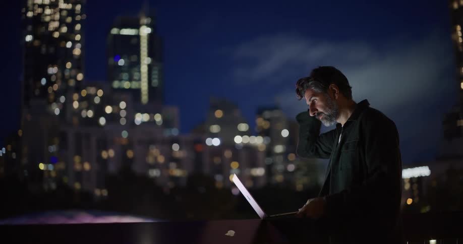 A man is standing outside looking at his laptop with the city as his backdrop