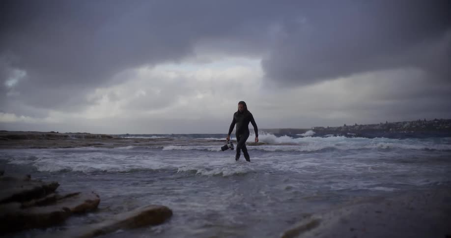 A man walks out of the ocean carrying a camera