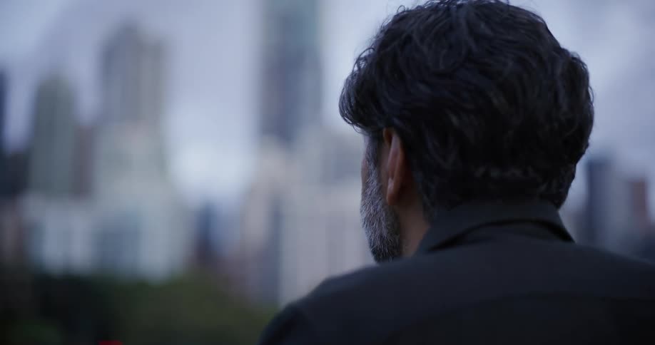 Bearded and black haired man looking around with buildings and trees on the background outdoors on an urban area during day time