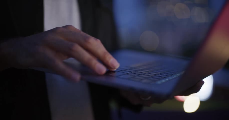 A persons hands are shown typing on a laptop keyboard at night with blurred lights in the background