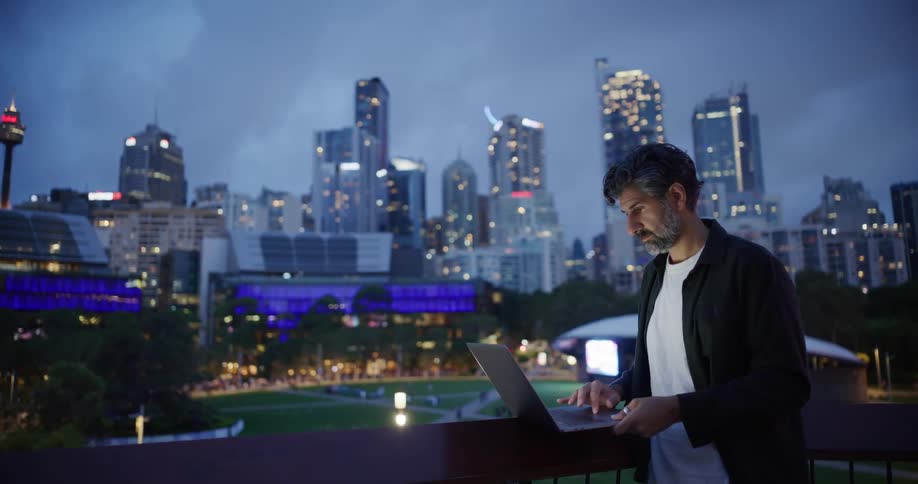 A person uses a laptop on a balcony overlooking a twilight cityscape with illuminated skyscrapers and a park below