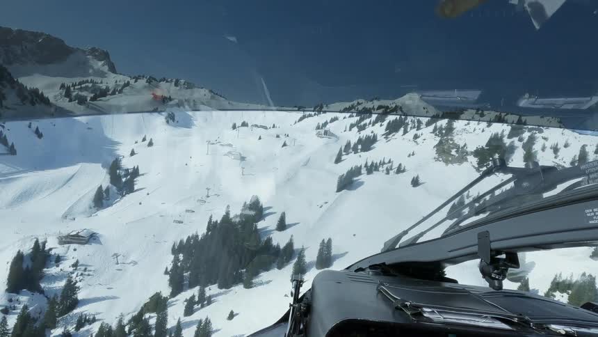 A view from inside a helicopter shows a snowy mountain slope with trees a few buildings and ski lifts below