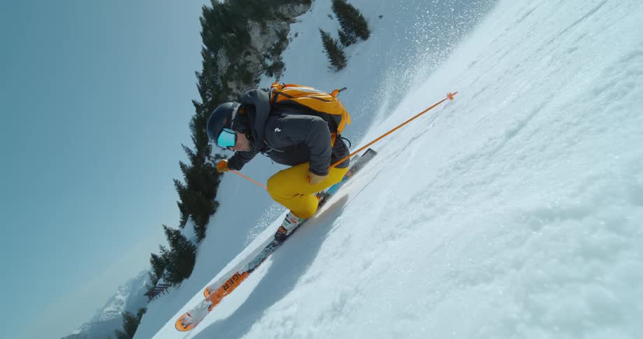 A skier in yellow pants and a dark jacket skiing down a steep snowy slope with trees and a mountain in the background