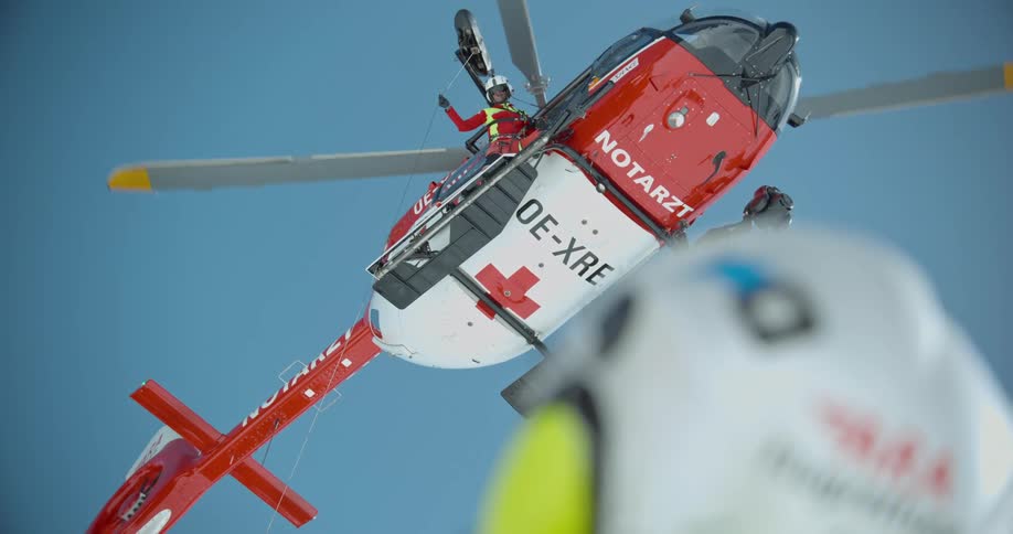 A red and white rescue helicopter hovers in the sky with a medical worker leaning out