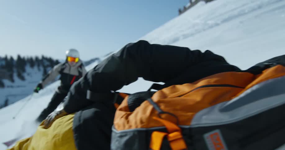 A man wearing ski gear fallen to the floor and another skiing towards him outdoors during day time