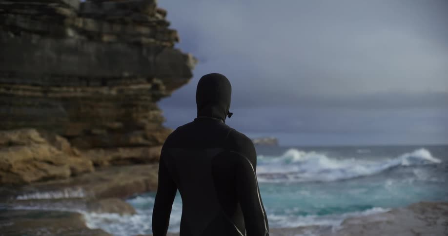 A person in a black wetsuit stands looking at a choppy ocean from a rocky coast under a cloudy sky