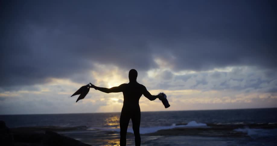 A silhouetted figure stands on a rocky shore at sunset holding fins and a camera raising his hands in celebration with a dramatic cloudy sky in the background