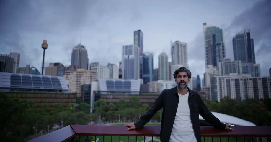 A man leans against the railing on the balcony in front of the city