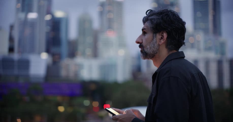 A man stands outdoors texting with a cityscape in the background at dusk