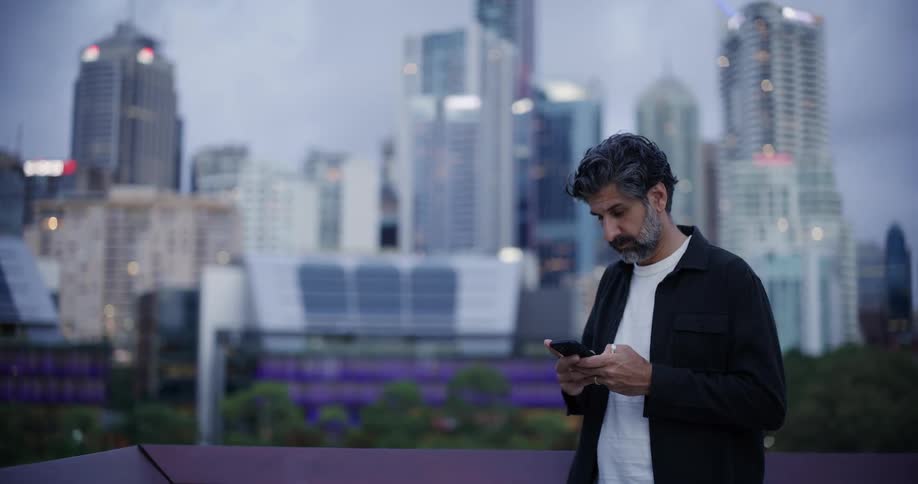 A man standing on a balcony looks at his phone then out at the city