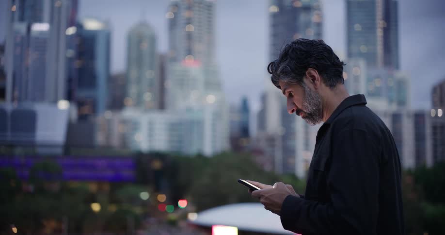 A man stands outdoors texting with a cityscape in the background at dusk