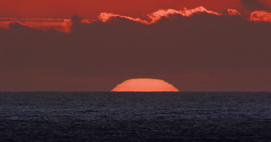 The sun rises over a calm ocean casting a fiery orange glow across the sky