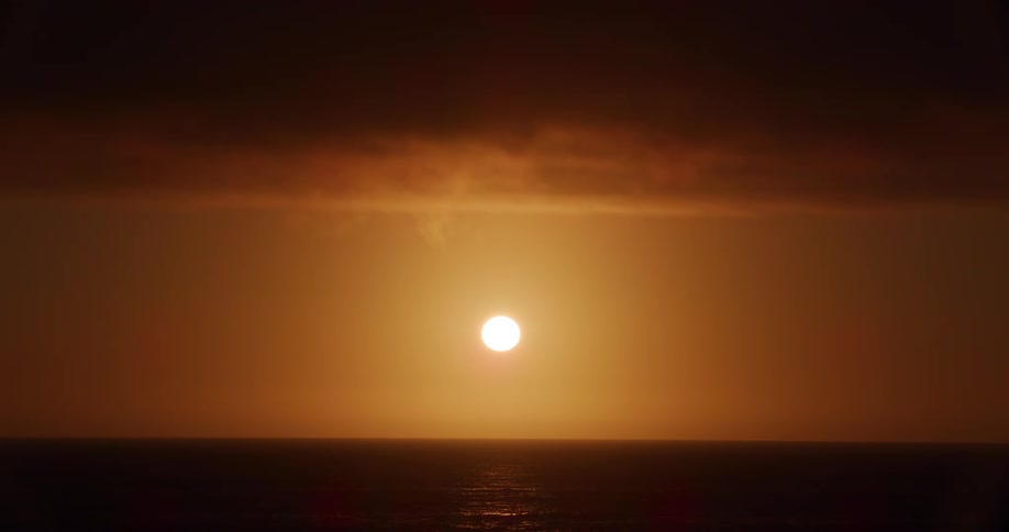 The sun sets over a calm ocean casting a warm orange glow across the sky