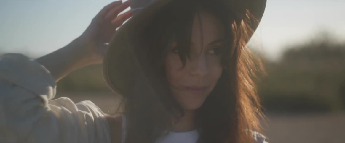A woman with long dark hair and a wide brimmed hat adjusting her hat while bathed in warm back lit sunlight