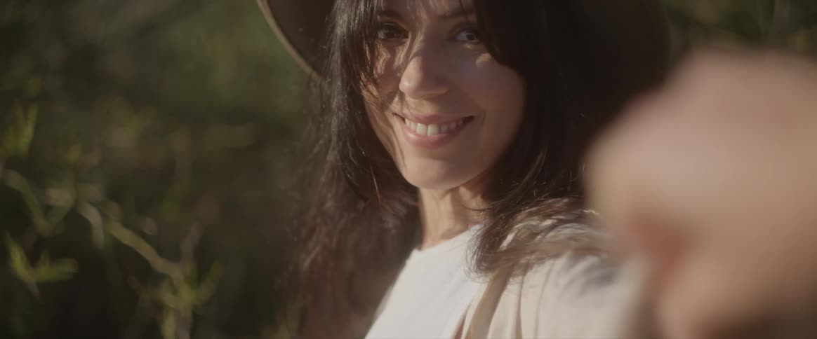 Dark haired woman wearing a hat holding a hand while she turns back and smiles outdoors during day time