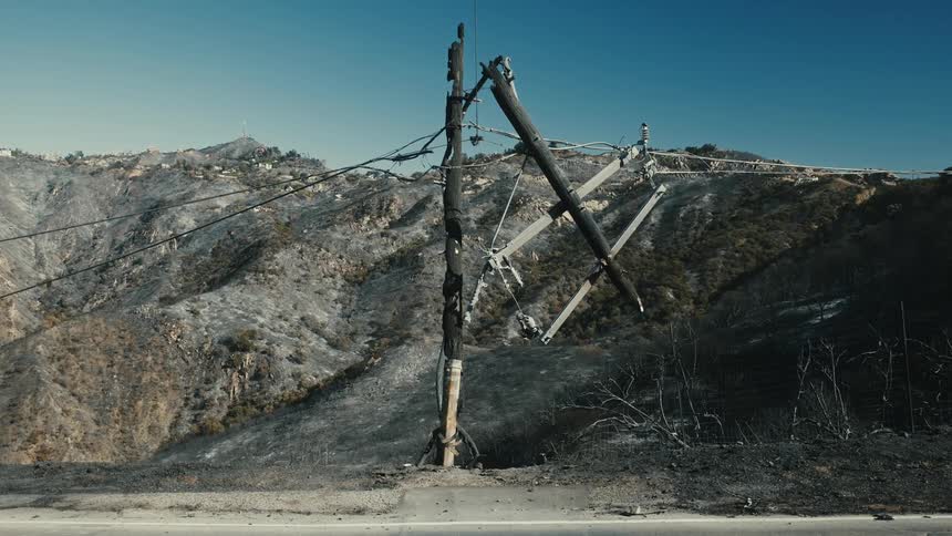 A charred utility pole leans precariously against a hillside power lines dangling amidst a landscape scorched by fire