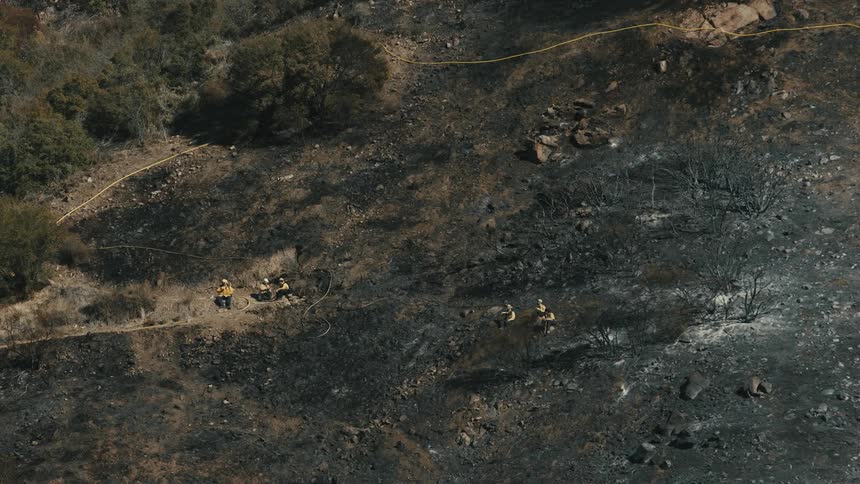 Four firefighters in yellow uniforms two grouped on a path and two further away working in a burned rocky landscape with a yellow hose visible
