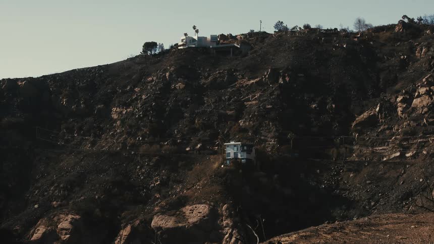 A hillside scorched black from a wildfire shows a small white house and a larger house higher up still standing amidst the devastation
