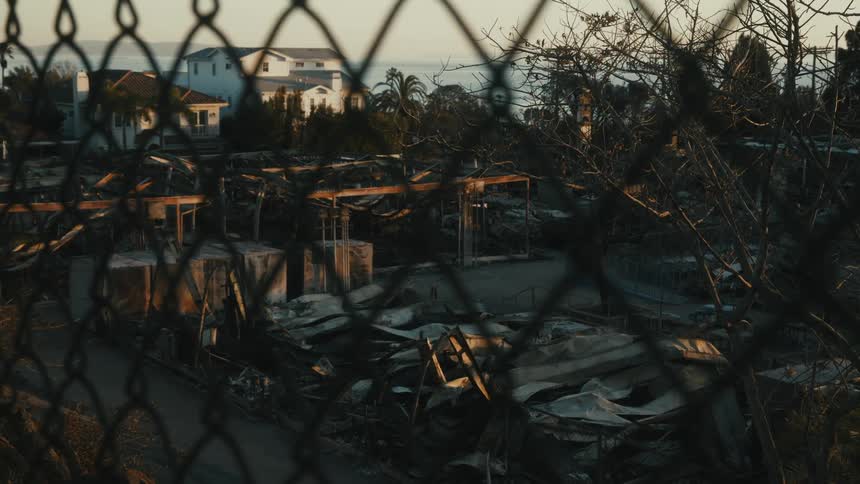 A view through a chainlink fence shows a dilapidated neighborhood with scattered debris and partially collapsed houses