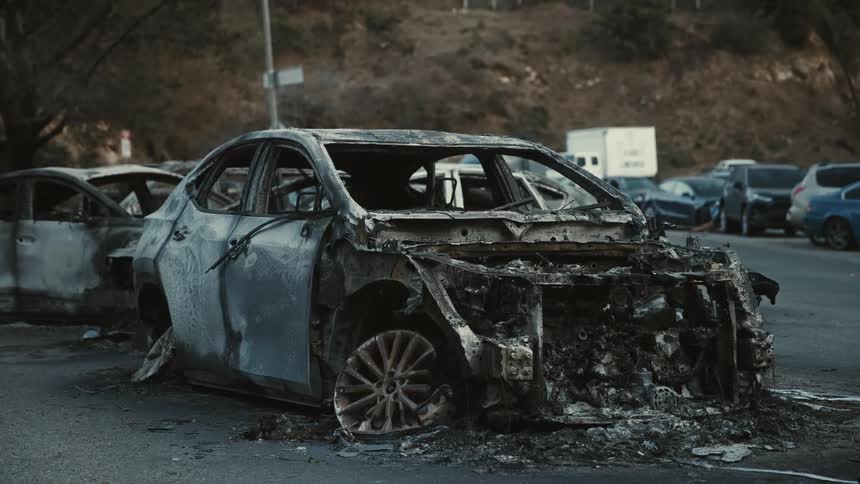 A severely fire damaged car sits in a parking lot among other damaged vehicles the aftermath of a wildfire