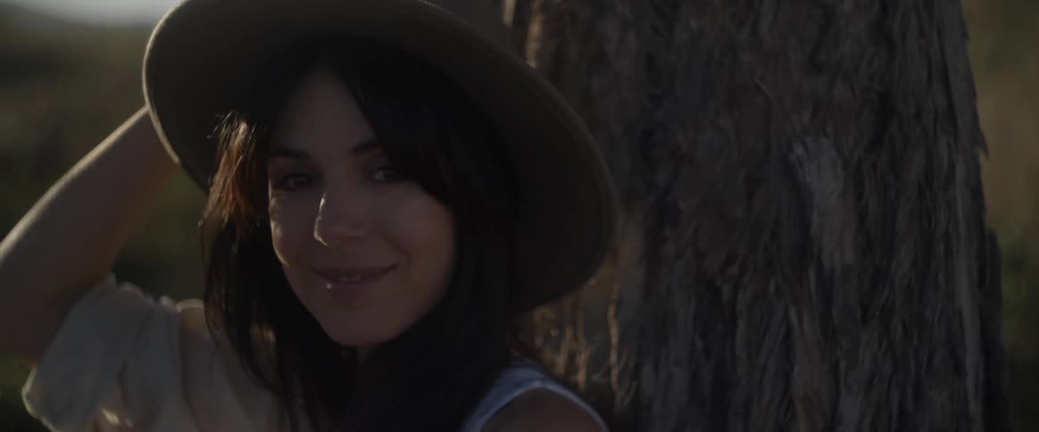 Portrait of a woman leaning against a tree smiling and putting on a sun hat