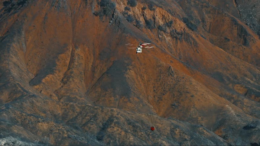 A firefighting helicopter is flying over rugged mountainous terrain with a red water bucket suspended below it