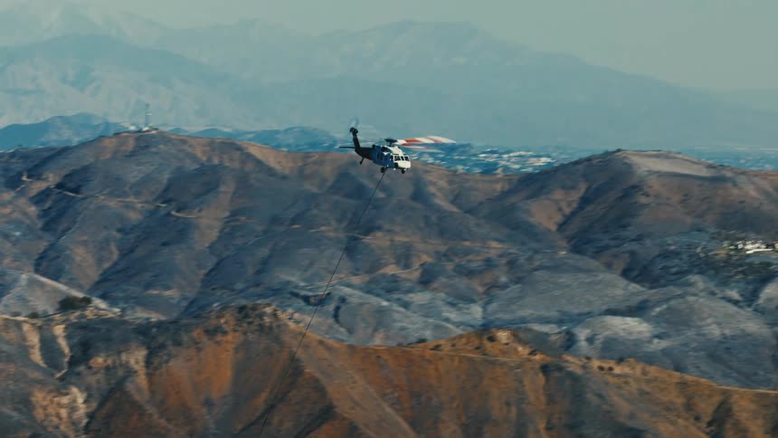 Rescue helicopter flies over a mountainous landscape carrying a cable as it passes through areas devastated by fire