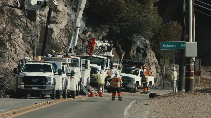 Team of workers wearing reflective vests performs road maintenance using trucks and equipment near a rocky outcrop