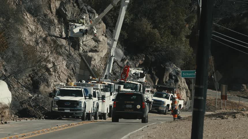 A line of utility trucks including a sheriffs vehicle is parked on a road by a rocky hillside where a worker is using a cherry picker