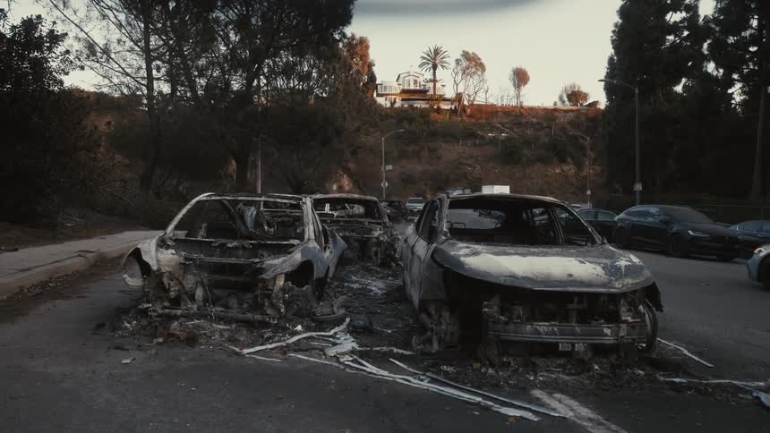Two fir damaged cars are shown parked on a street with other vehicles and a hillside house visible in the background