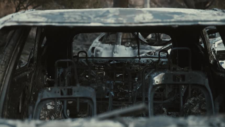 The interior of a fire ravaged car is shown revealing severely burned seats and dashboard with other damaged vehicles visible in the background