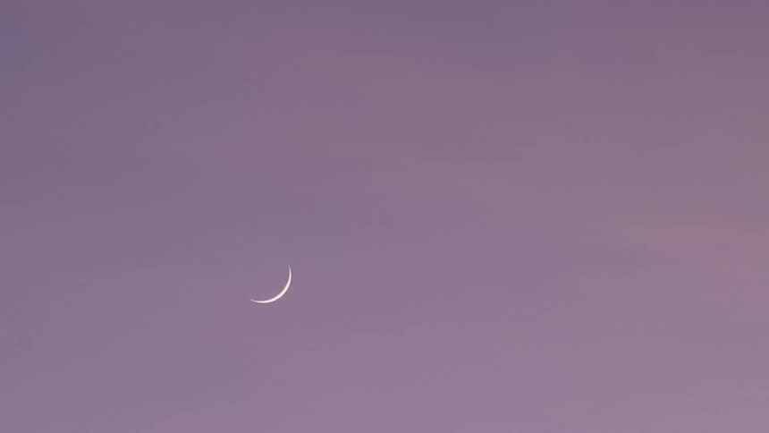 A crescent moon hangs faintly in a pale lavenderpink sky