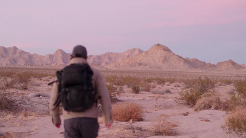 A slightly out of focus person wearing a cap and a dark backpack is walking away from the viewer on a sandy trail in a desert landscape with mountains in the background under a pinkish sky