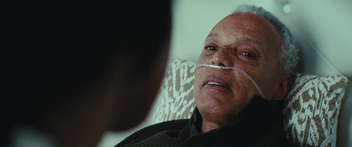 An elderly person with a nasal cannula is lying on a bed talking to another person