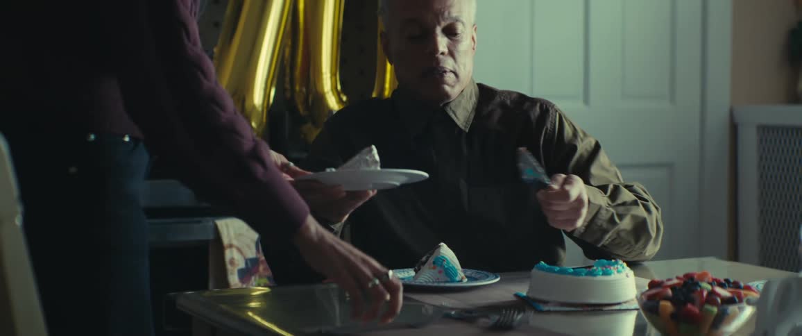 An elderly person is cutting a slice of birthday cake on a plate for another person in a room decorated with balloons