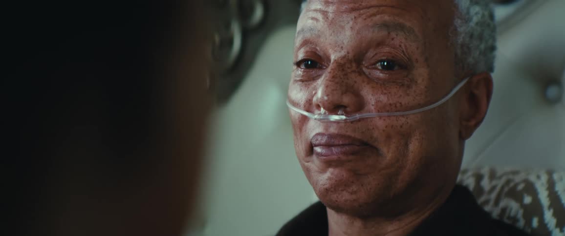 An elderly person with a nasal cannula is lying on a bed talking to another person