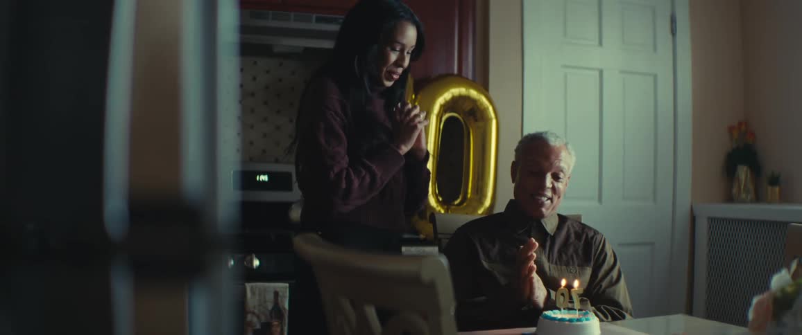 An elderly person celebrates his birthday with a small cake with two lit candles and a large gold balloon shaped like the number 70 in the background