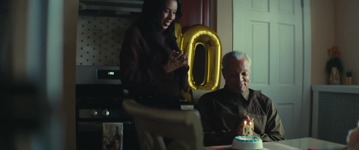 Woman placing a birthday cake with lit candles for an elderly man who is sitting at the table happily
