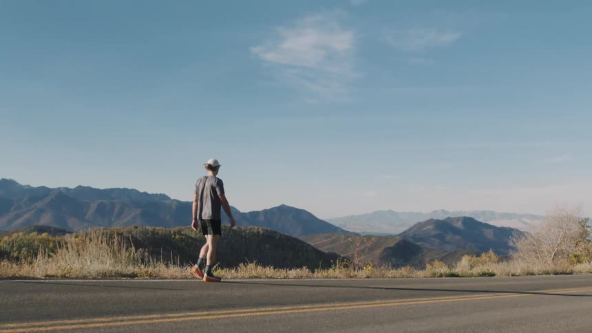 Man walks along the edge of a highway passing through hilly terrain on a sunny day
