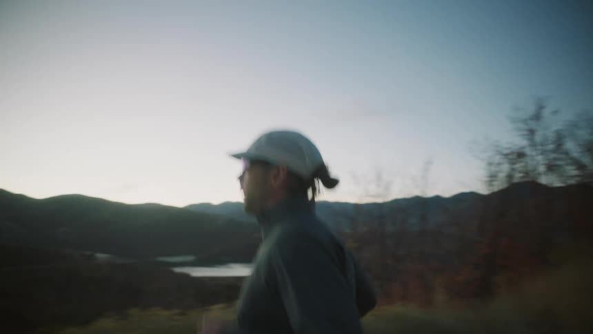 A person wearing a white cap and dark clothing is jogging along a road with a scenic view of rolling hills and a distant lake at dusk