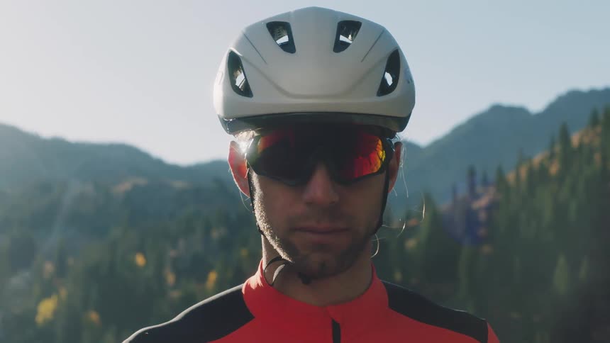 A cyclist wearing a white helmet and redtinted sunglasses looking directly forward with a mountainous forested backdrop on a sunny day