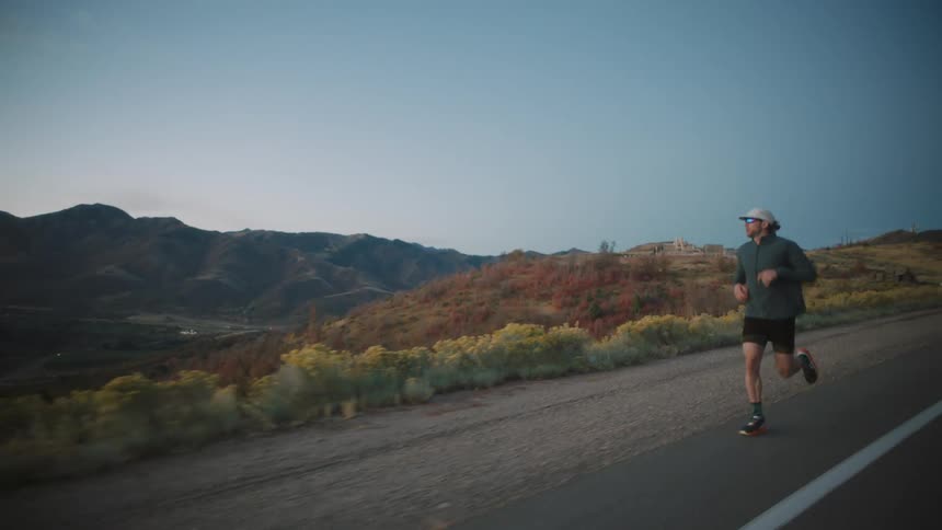 A man wearing a gray long sleeve jacket black shorts a white cap and sunglasses runs along a paved road with mountains in the background under a twilight sky